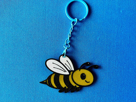 A Bee Keyring laying on a blue tablecloth. The Bee keyring is made of PLA plastic and is coloured Black, Yellow and White, with a coloured stainless steel key chain.