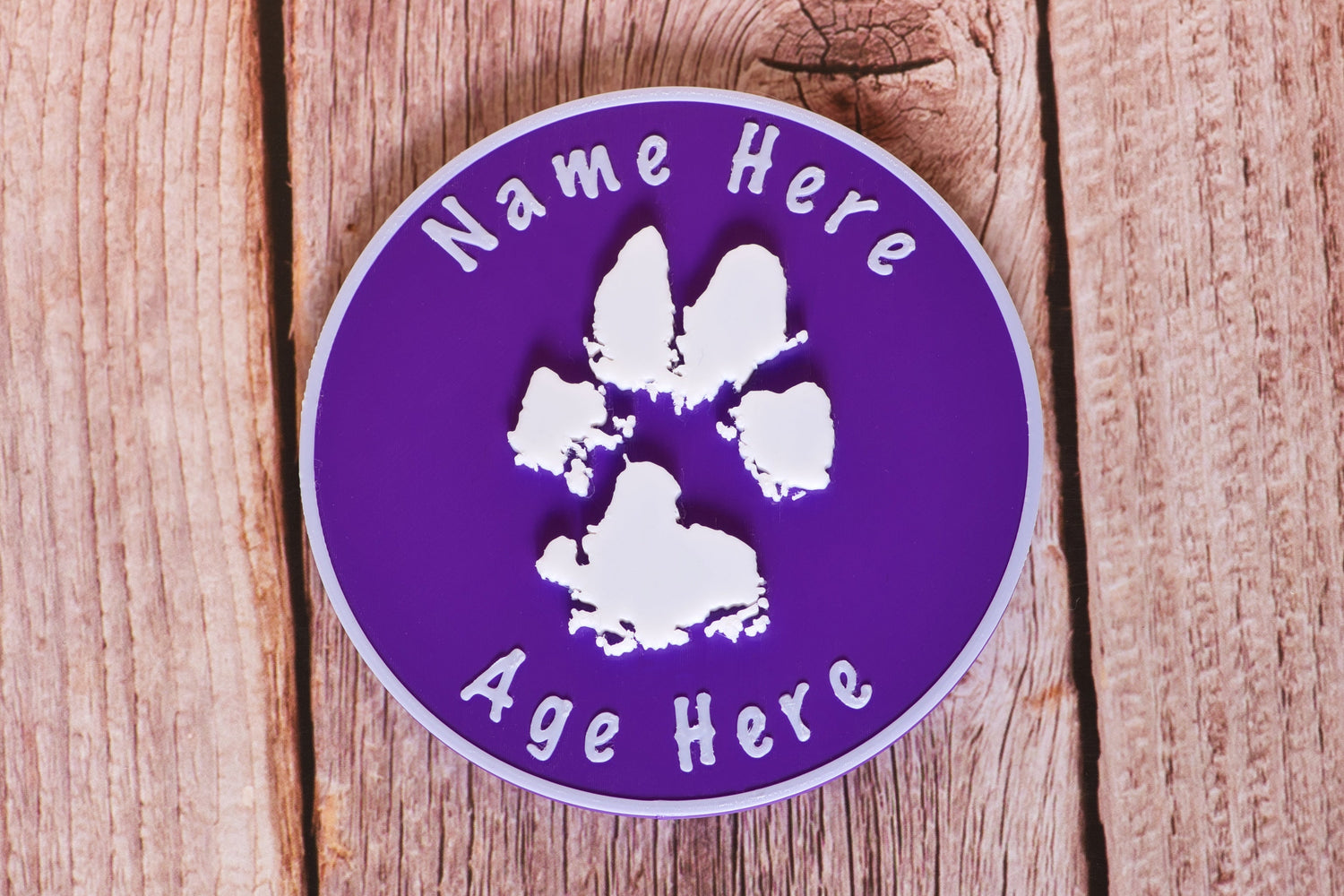 Round Paw Print Standing Plaque laying on a wooden surface. The plaque features a white 3D Paw Print and text on a purple background