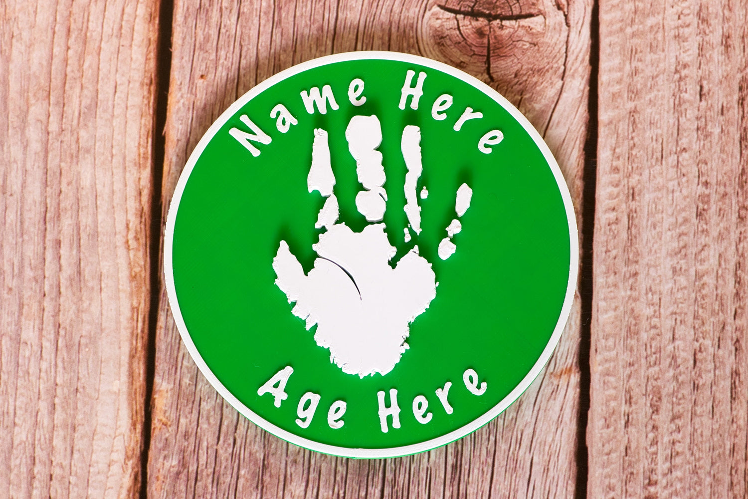 3D Round Child Handprint Standing Plaque laying on a wooden surface. The Plaque features a white handprint and text on a green background