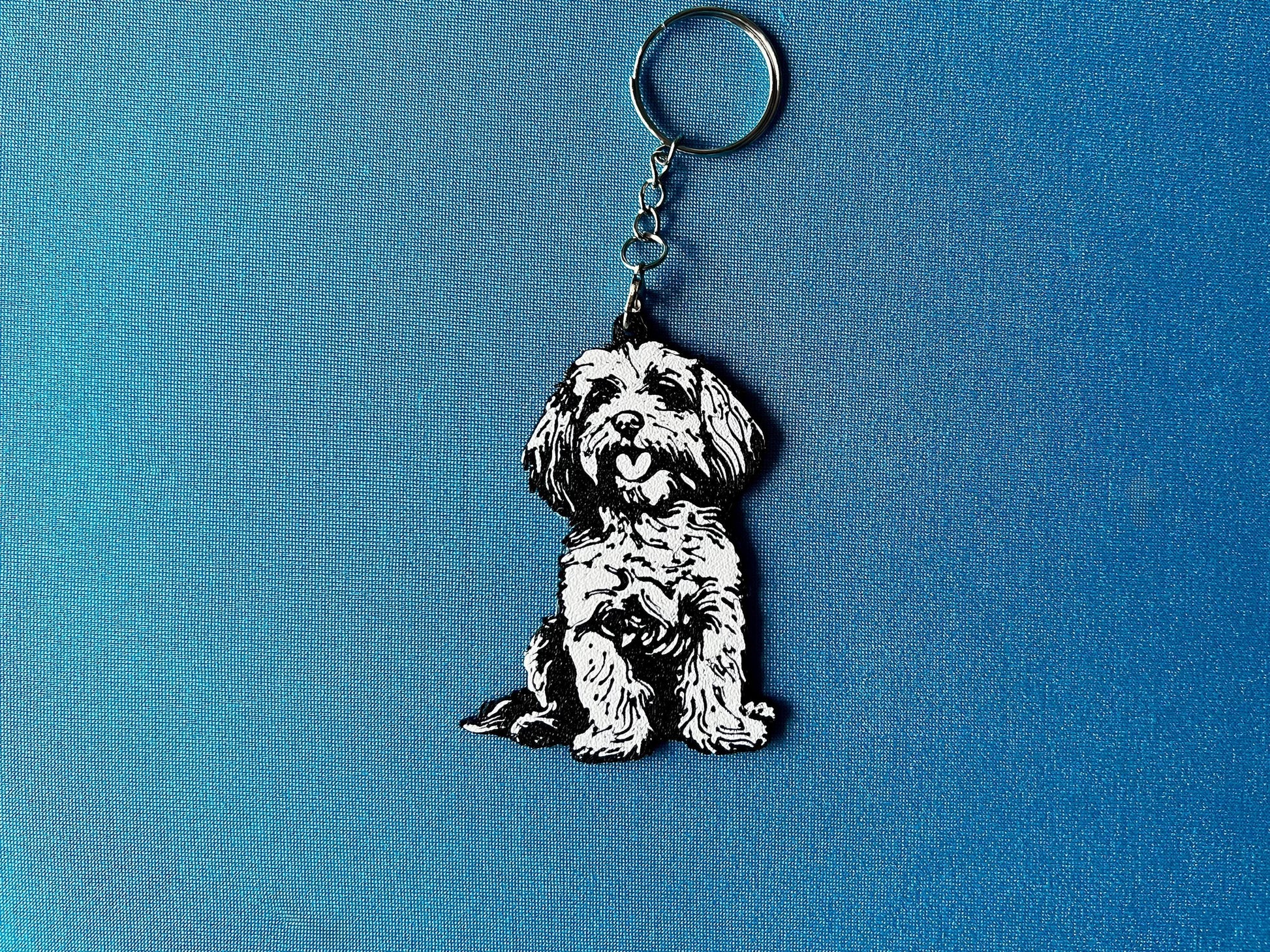 Cockapoo Silhouette Keyring laying on a blue table cloth. The keyring features a white background with a black silhouette of a Cockerpoo.