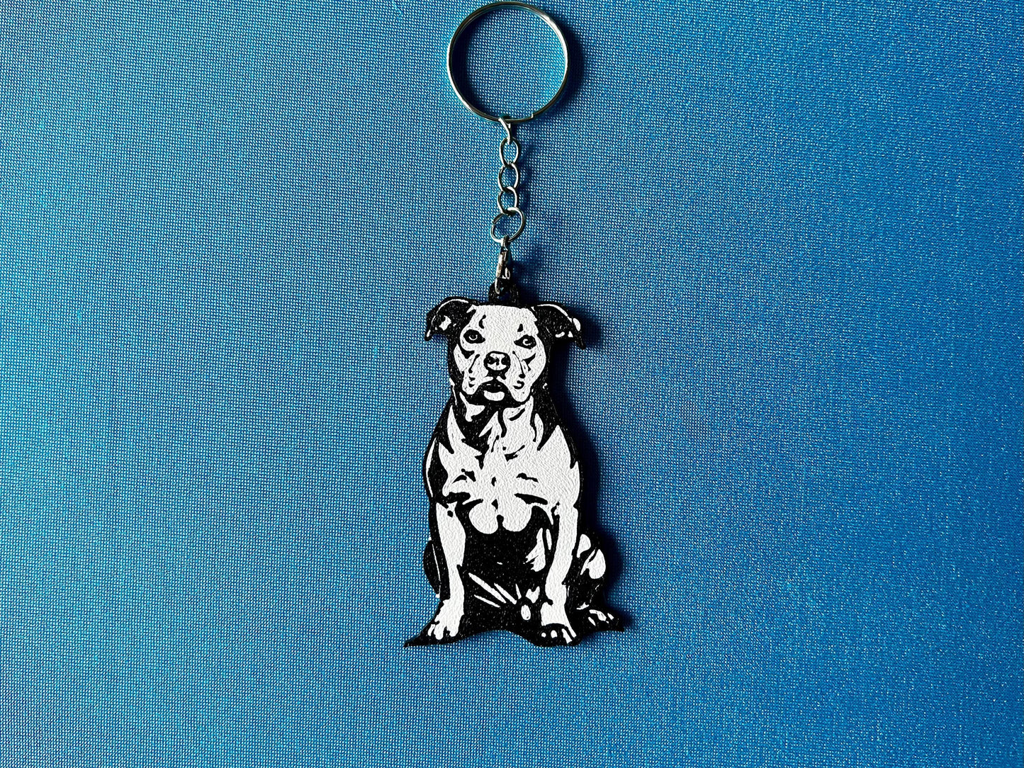 Staffordshire Bull Terrier Silhouette Keyring laying on a blue table cloth. The keyring features a white background with a black silhouette of a Staffordshire Bull Terrier.