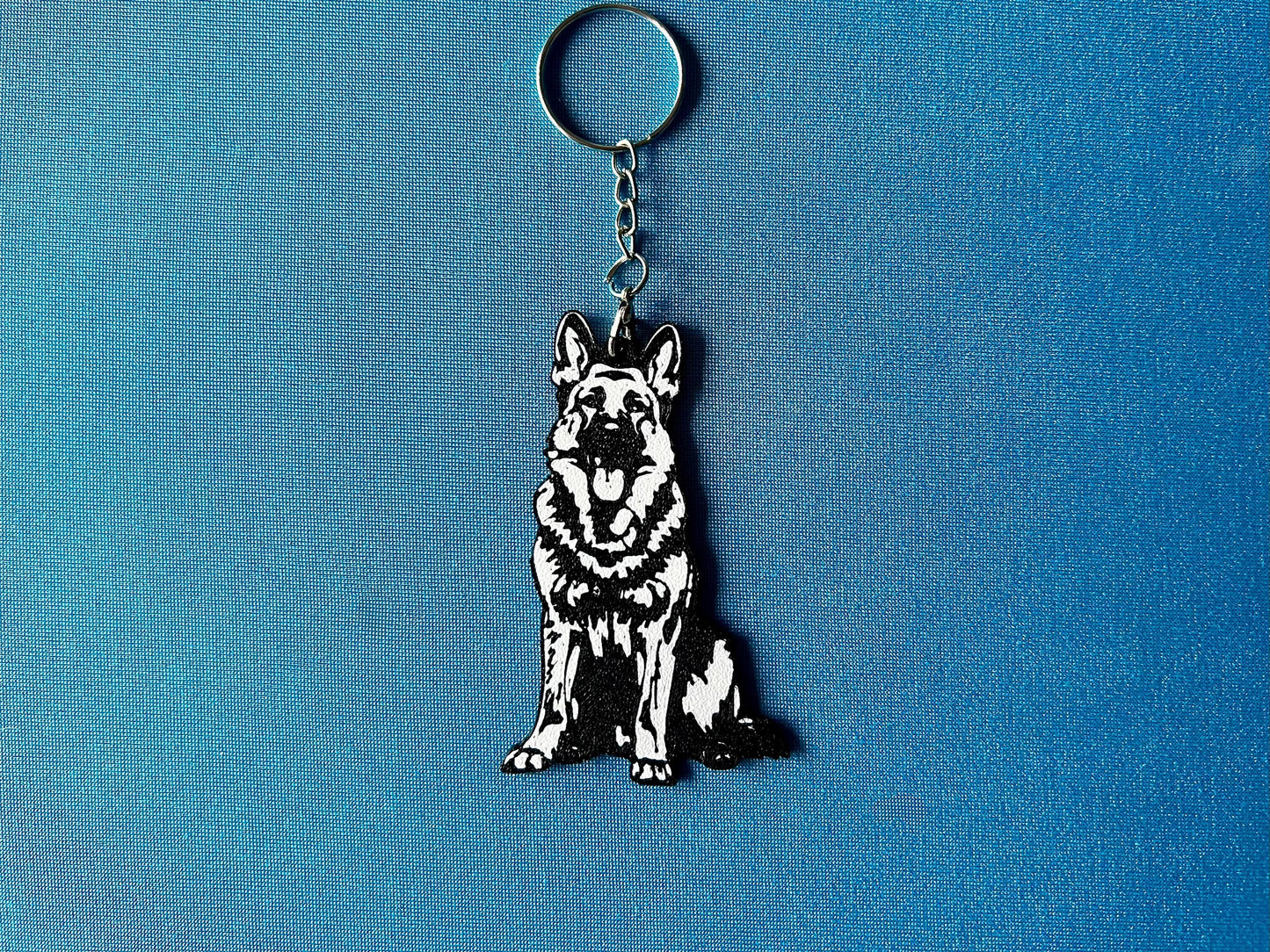 German Shepherd Silhouette Keyring laying on a blue table cloth. The keyring features a white background with a black silhouette of a German Shepherd.