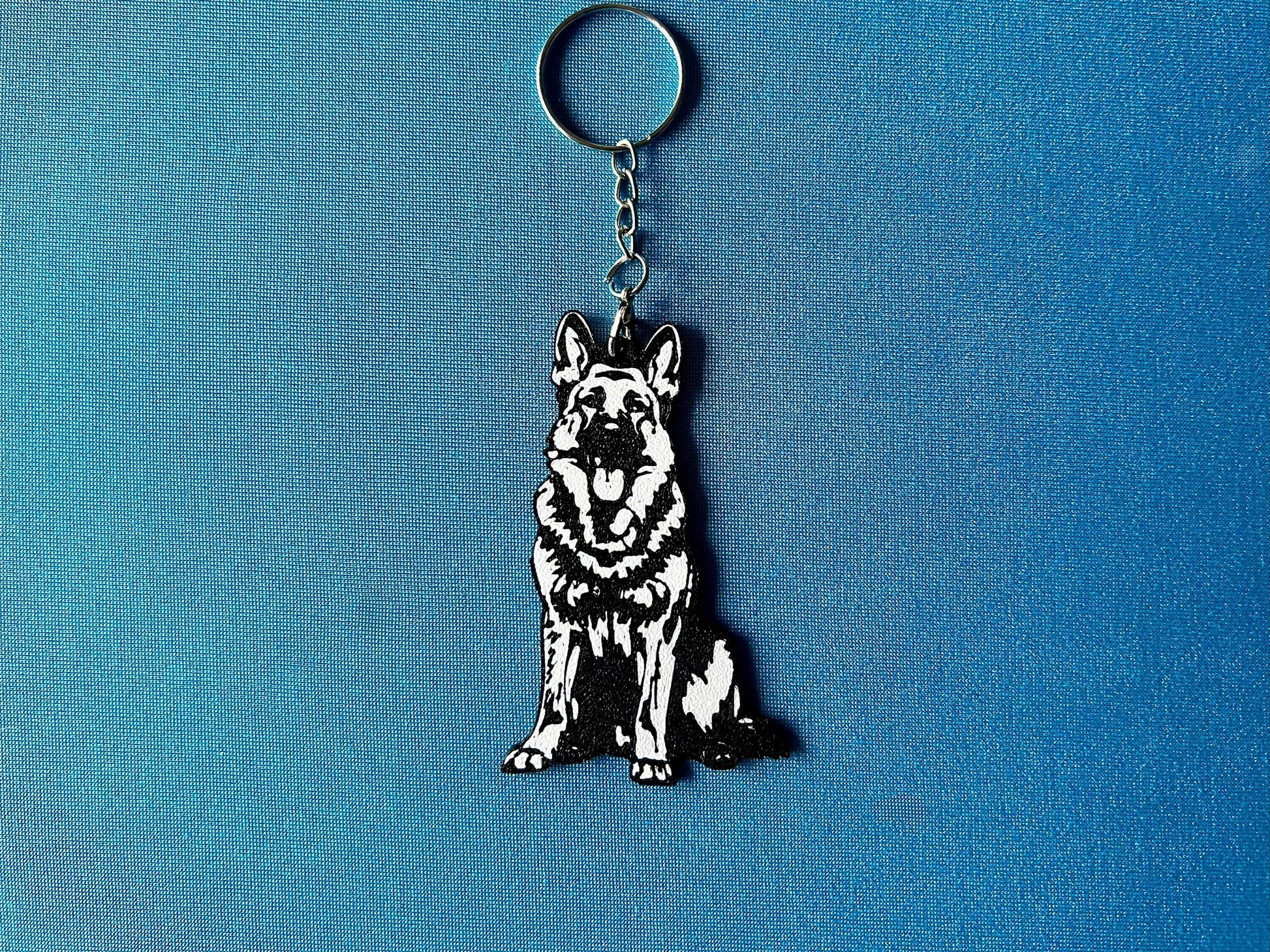 German Shepherd Silhouette Keyring laying on a blue table cloth. The keyring features a white background with a black silhouette of a German Shepherd.