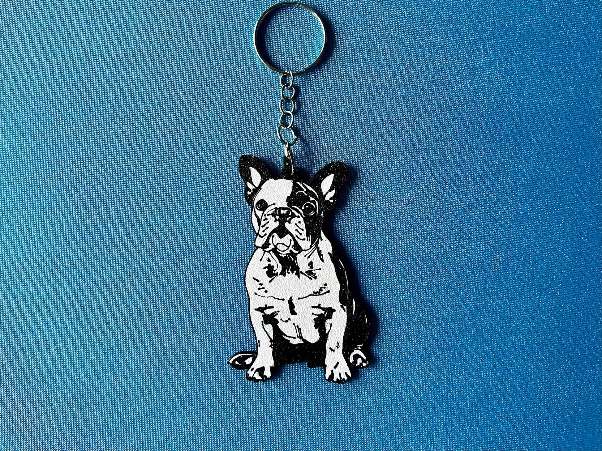 French Bulldog Silhouette Keyring laying on a blue table cloth. The keyring features a white background with a black silhouette of a French Bulldog.