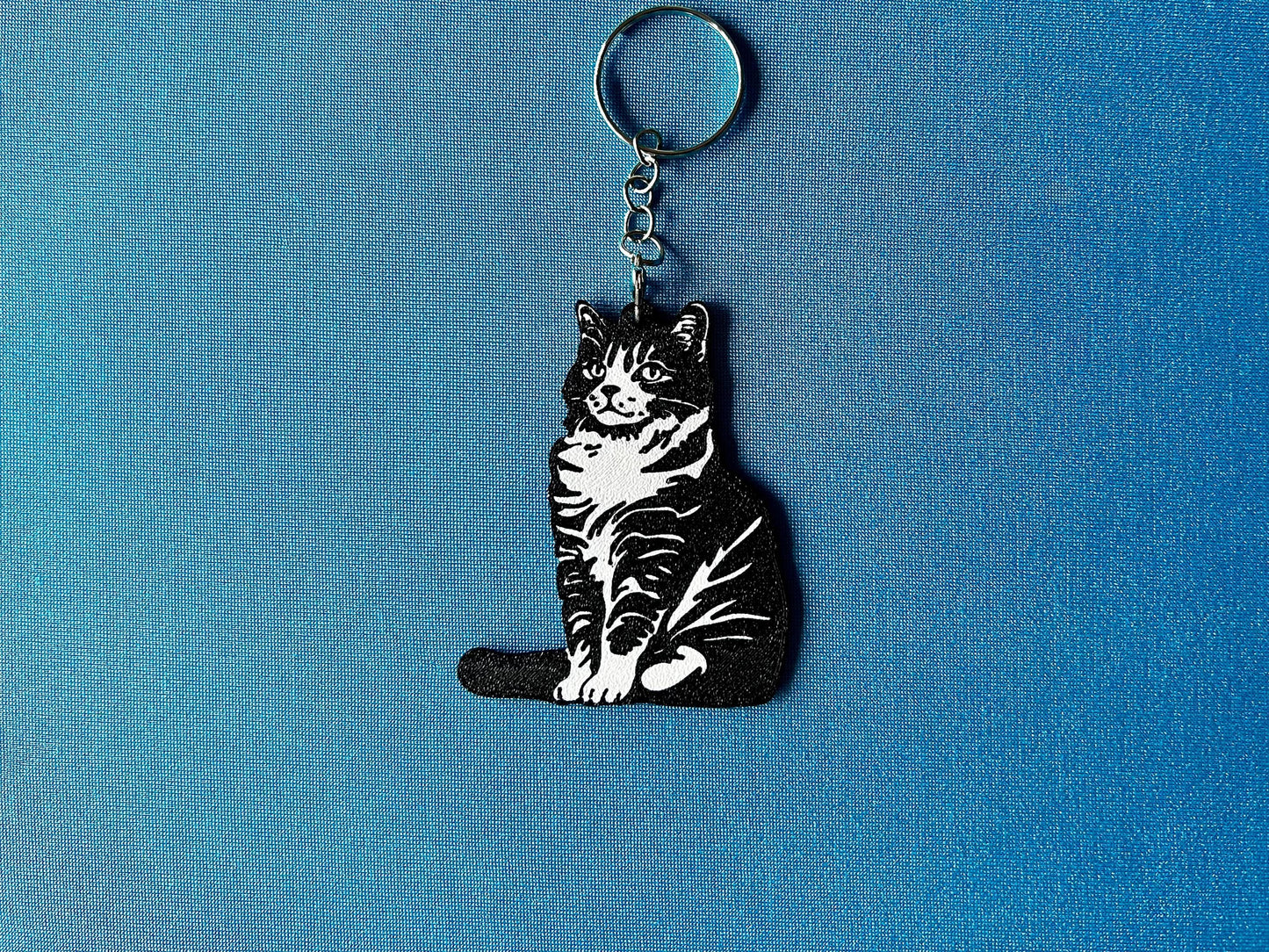 American Bobtail Silhouette Keyring laying on a blue table cloth. The keyring features a white background with a black silhouette of a American Bobtail.