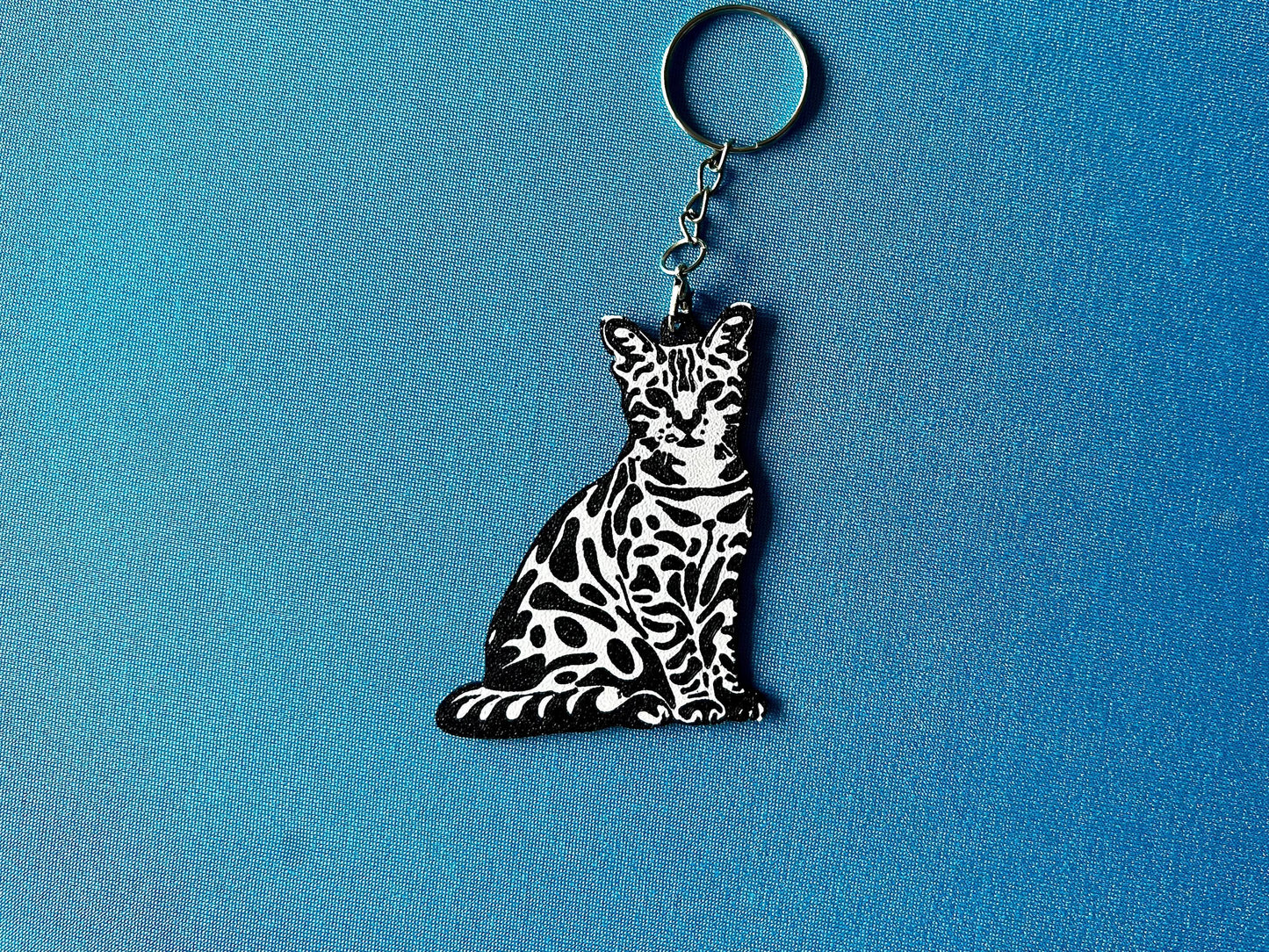 Bengal Silhouette Keyring laying on a blue table cloth. The keyring features a white background with a black silhouette of a Bengal.