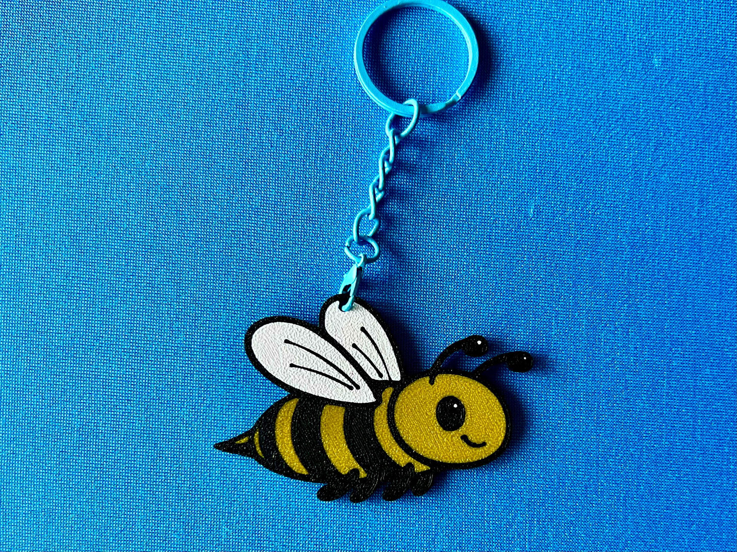 A Bee Keyring laying on a blue tablecloth. The Bee keyring is made of PLA plastic and is coloured Black, Yellow and White, with a coloured stainless steel key chain.