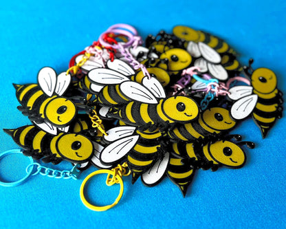 A pile of Bee Keyrings laying on a blue tablecloth. The Bee keyrings are made of PLA plastic and are coloured Black, Yellow and White, with a coloured stainless steel key chain.