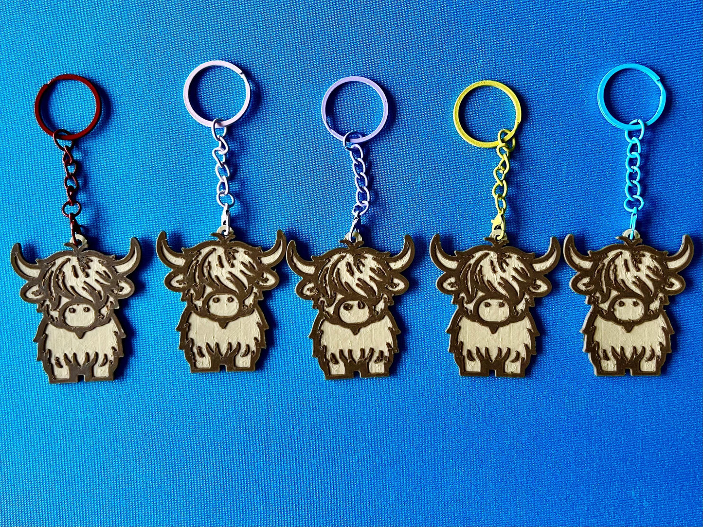 Highland Cow Keyrings in a line, laying on a blue tablecloth. The Highland Cow keyrings are made of Wood PLA and are Maple and Walnut coloured, with a coloured stainless steel key chain.