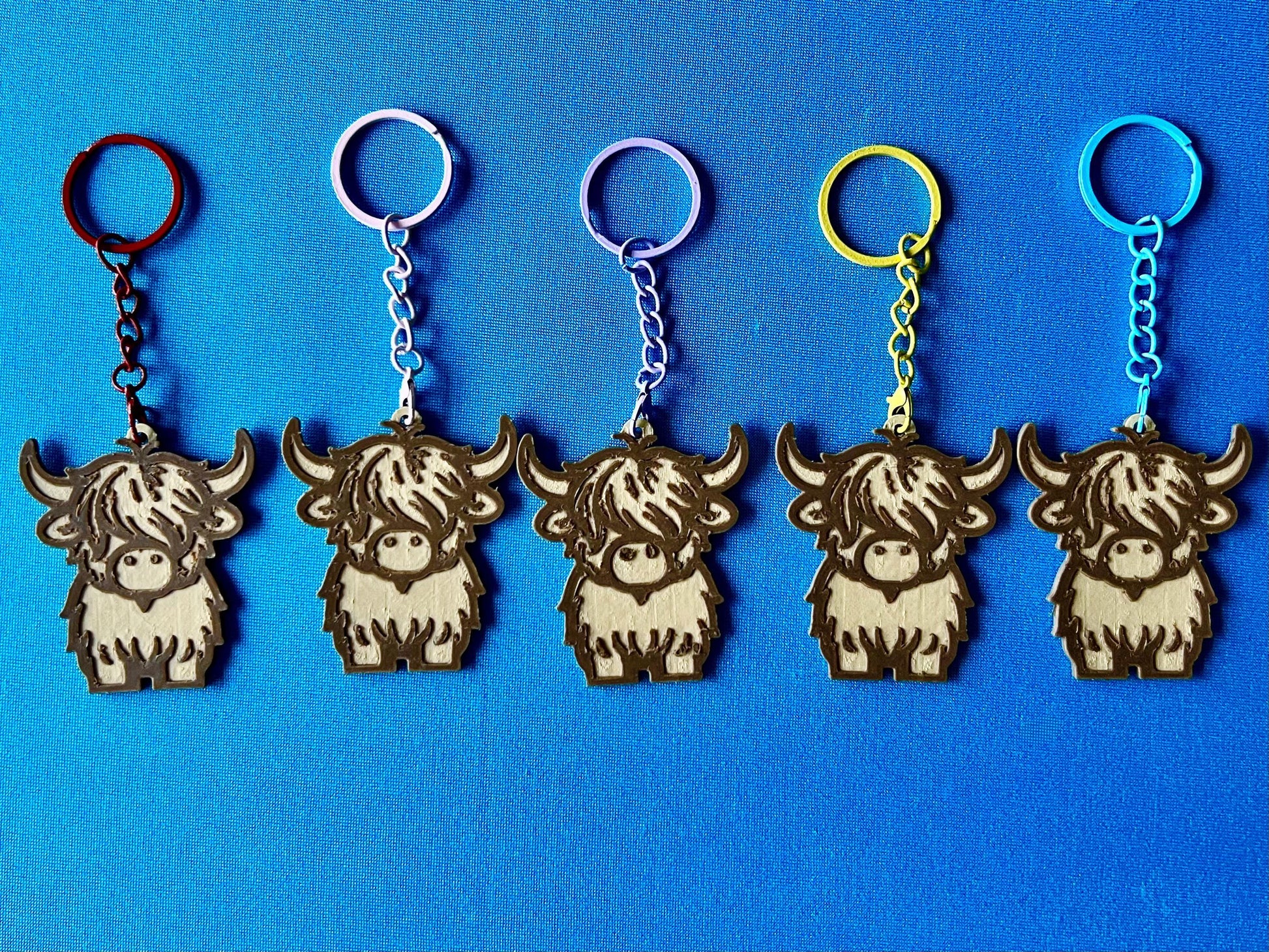 Highland Cow Keyrings in a line, laying on a blue tablecloth. The Highland Cow keyrings are made of Wood PLA and are Maple and Walnut coloured, with a coloured stainless steel key chain.