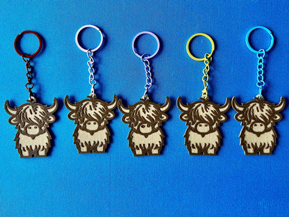 Highland Cow Keyrings in a line, laying on a blue tablecloth. The Highland Cow keyrings are made of Wood PLA and are Maple and Walnut coloured, with a coloured stainless steel key chain.