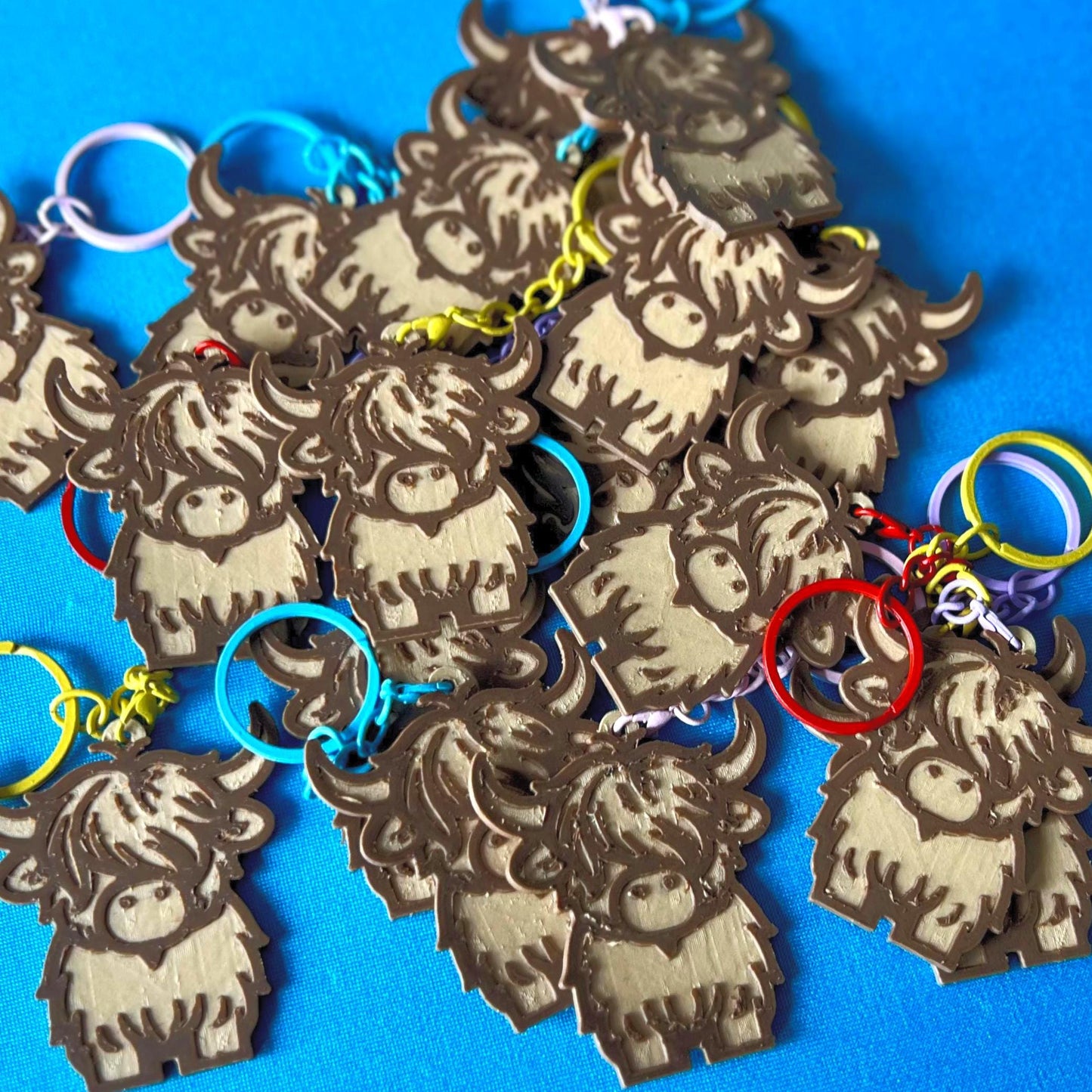 A pile of Highland Cow Keyrings laying on a blue tablecloth. The Highland Cow keyrings are made of Wood PLA and are Maple and Walnut coloured, with a coloured stainless steel key chain.
