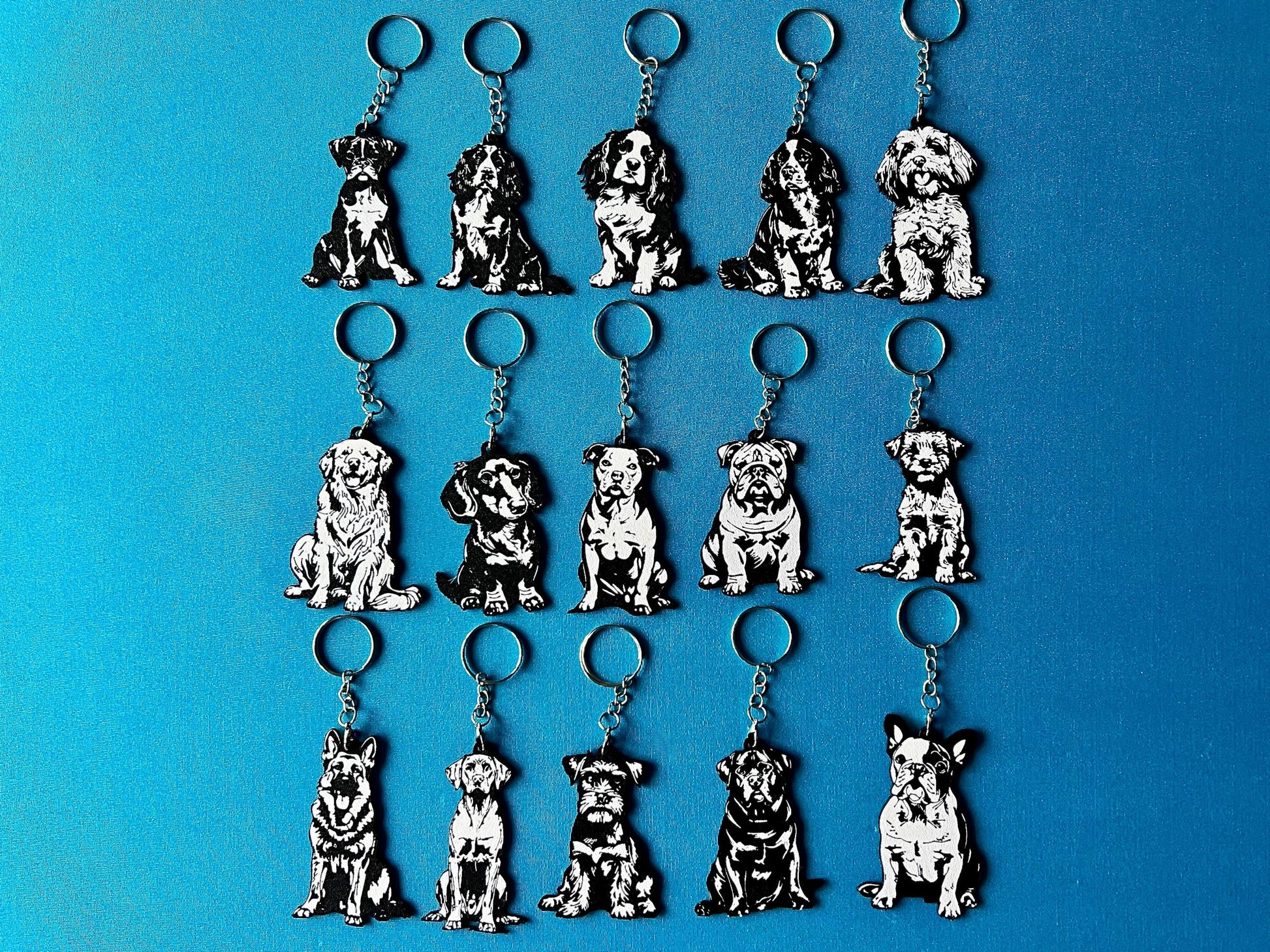 15 Dog breed Silhouette Keyrings laying on a blue table cloth. The keyrings feature a white background with a black silhouette of a dog.