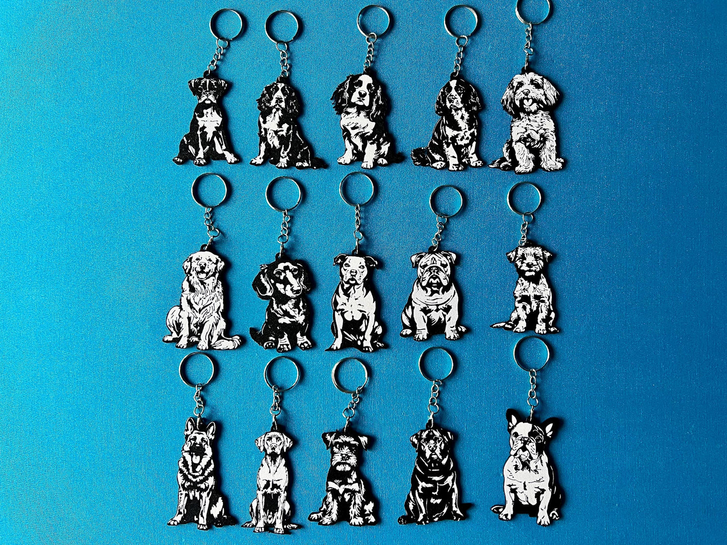 15 Dog breed Silhouette Keyrings laying on a blue table cloth. The keyrings feature a white background with a black silhouette of a dog.