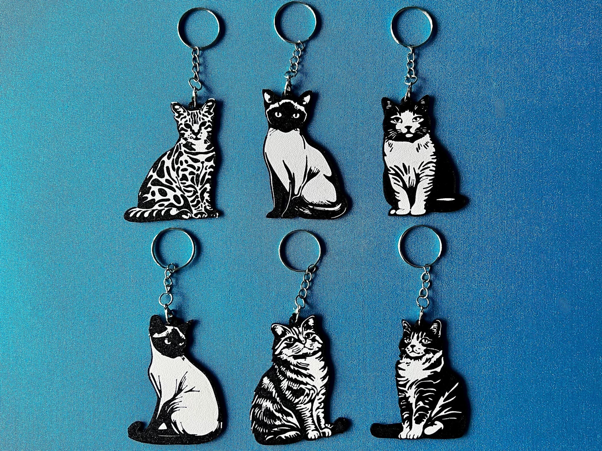 6 Cat breed Silhouette Keyrings laying on a blue table cloth. The keyrings feature a white background with a black silhouette of a cat.