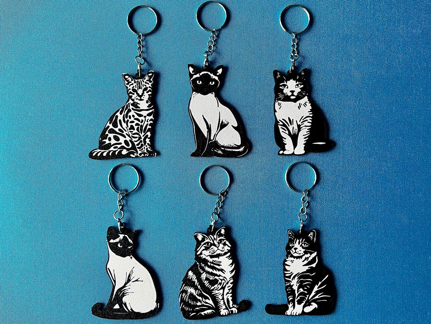 6 Cat breed Silhouette Keyrings laying on a blue table cloth. The keyrings feature a white background with a black silhouette of a cat.