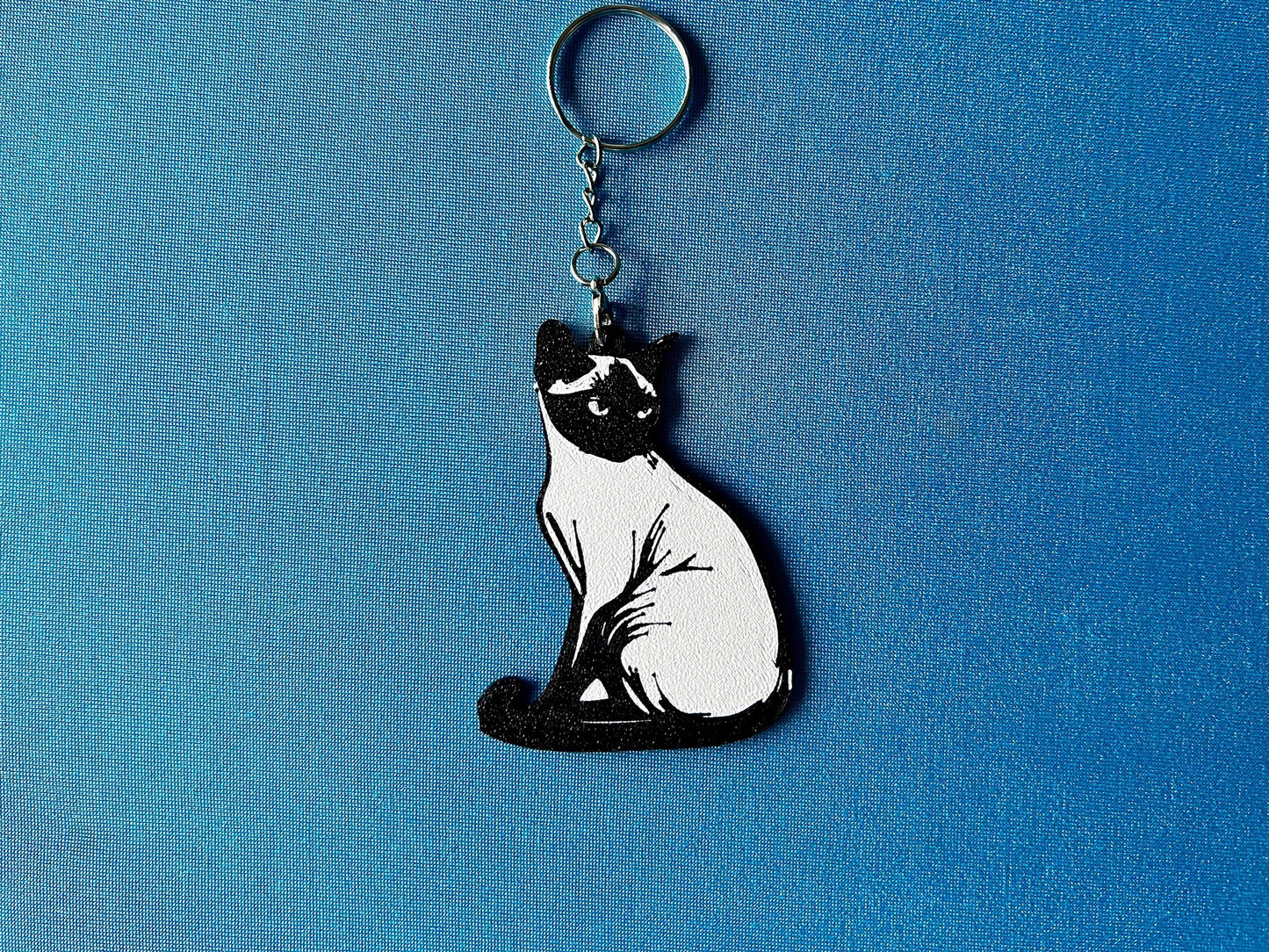 Burmese Silhouette Keyring laying on a blue table cloth. The keyring features a white background with a black silhouette of a Burmese.