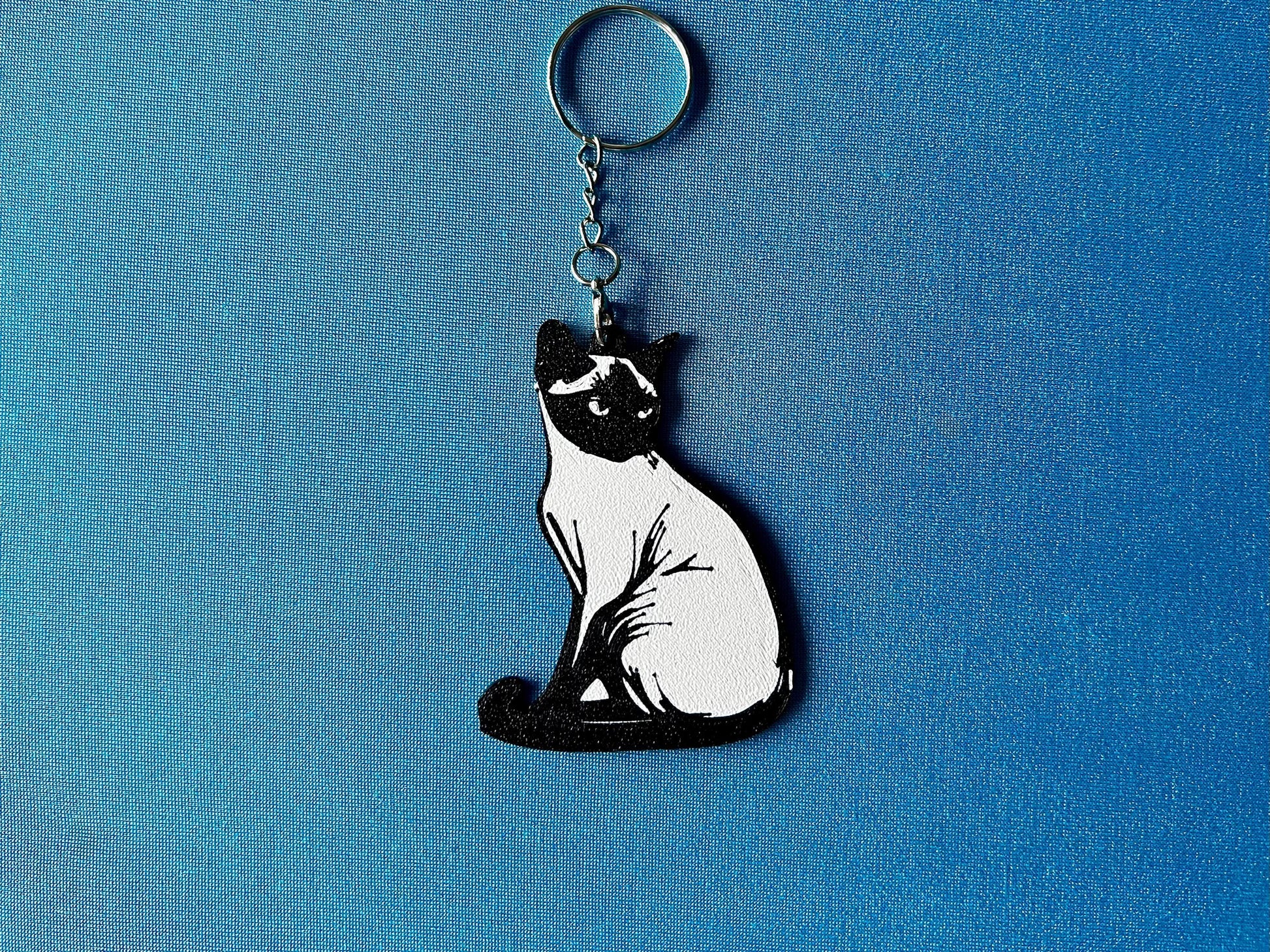 Burmese Silhouette Keyring laying on a blue table cloth. The keyring features a white background with a black silhouette of a Burmese.