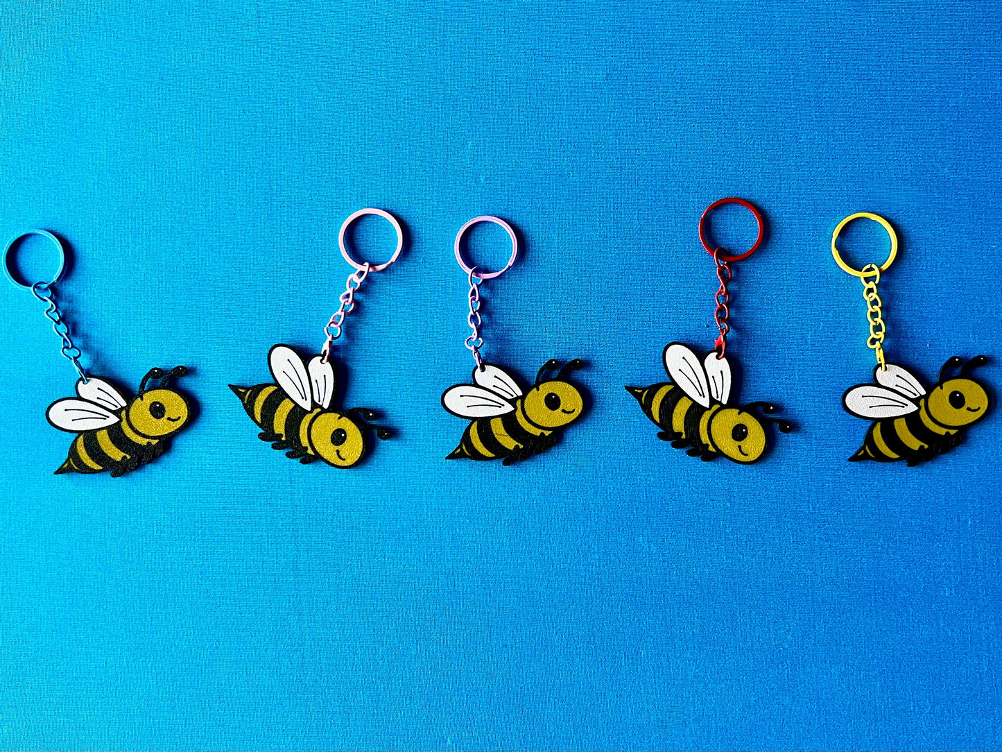 Bee Keyrings in a line, laying on a blue tablecloth. The Bee keyrings are made of PLA plastic and are coloured Black, Yellow and White, with a coloured stainless steel key chain.