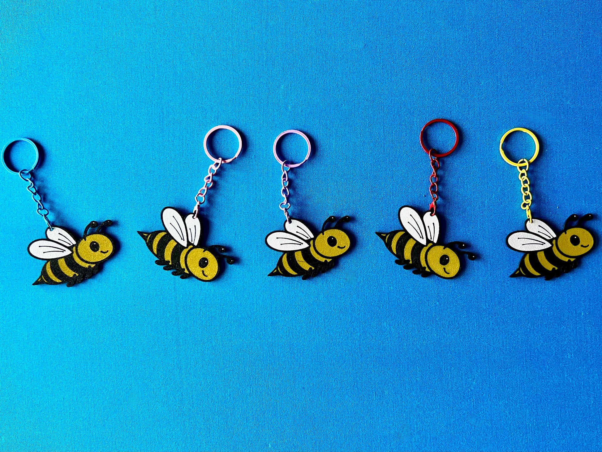 Bee Keyrings in a line, laying on a blue tablecloth. The Bee keyrings are made of PLA plastic and are coloured Black, Yellow and White, with a coloured stainless steel key chain.