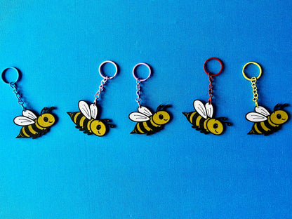 Bee Keyrings in a line, laying on a blue tablecloth. The Bee keyrings are made of PLA plastic and are coloured Black, Yellow and White, with a coloured stainless steel key chain.