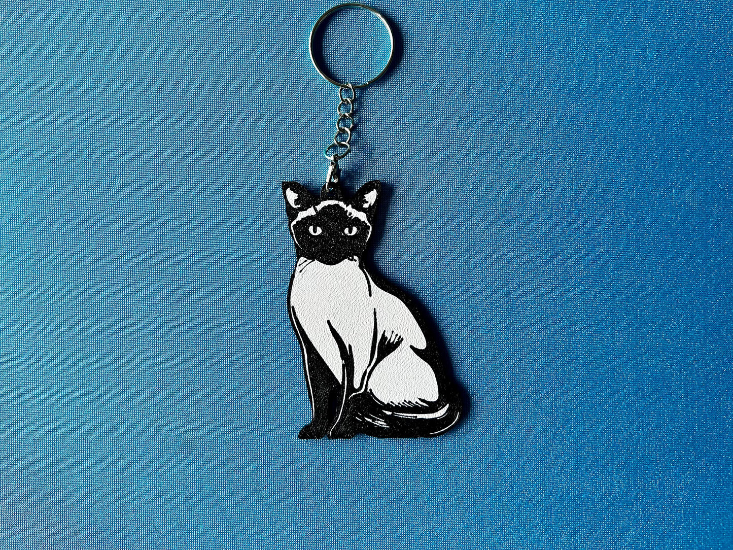Siamese Silhouette Keyring laying on a blue table cloth. The keyring features a white background with a black silhouette of a Siamese.