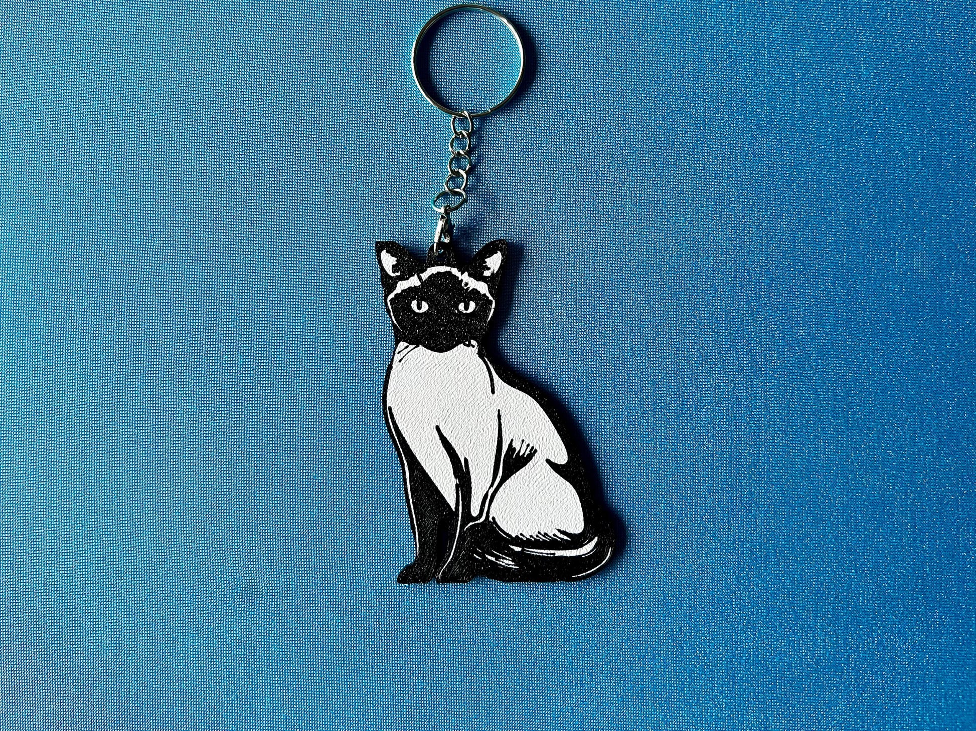 Siamese Silhouette Keyring laying on a blue table cloth. The keyring features a white background with a black silhouette of a Siamese.