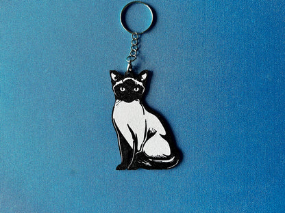 Siamese Silhouette Keyring laying on a blue table cloth. The keyring features a white background with a black silhouette of a Siamese.