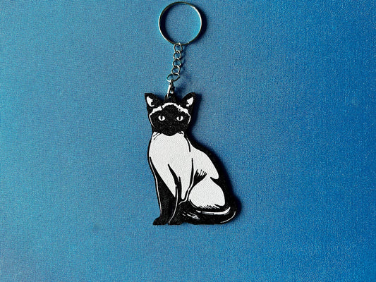Siamese Silhouette Keyring laying on a blue table cloth. The keyring features a white background with a black silhouette of a Siamese.