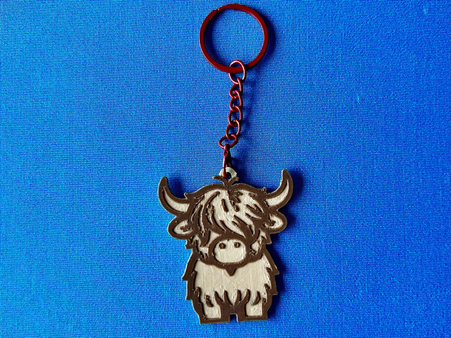 A Highland Cow Keyring laying on a blue tablecloth. The Highland Cow keyring is made of Wood PLA and is coloured in two tones of wood (Walnut and Maple), with a coloured stainless steel key chain.