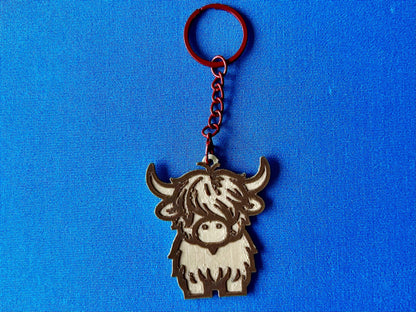 A Highland Cow Keyring laying on a blue tablecloth. The Highland Cow keyring is made of Wood PLA and is coloured in two tones of wood (Walnut and Maple), with a coloured stainless steel key chain.