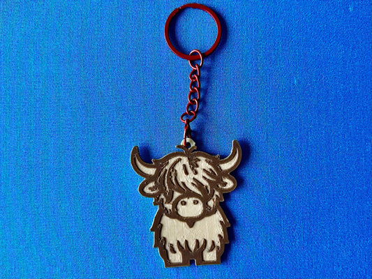 A Highland Cow Keyring laying on a blue tablecloth. The Highland Cow keyring is made of Wood PLA and is coloured in two tones of wood (Walnut and Maple), with a coloured stainless steel key chain.